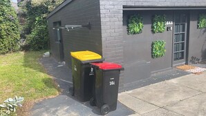 Two trash bins with yellow and red lids, labeled '164', placed outside a building with a grey brick facade and green decorative elements. - Mentone Compact Backyard Cabin Beachside (Parkdale)