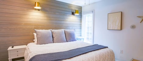 A bedroom with a wooden headboard, a bed with a white quilt, a dark blue throw blanket, a nightstand with a lamp, and a ceiling fan.
