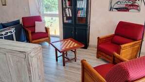 Ein gemütliches Wohnzimmer mit Couchtisch aus Holz, roten Polstersesseln, einem Bücherregal und Blick ins Freie.