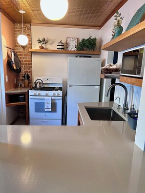A compact kitchen with a built-in oven, a refrigerator, a microwave, and a sink. There are hanging pots and pans, a kettle, and a shelf with decorative items. - Quaint sunny cottage that rests in the historical neighborhood of Billings (Billings)