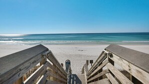 Une promenade en bois mène à une plage de sable fin, sous un ciel bleu et dégagé.