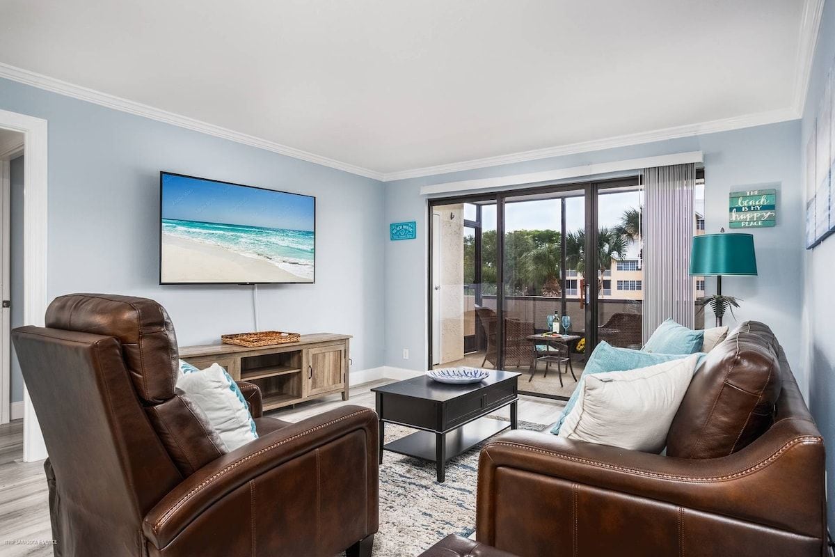 A living room with leather sofas, a flat-screen TV, a coffee table, and a view of the beach through a sliding glass door.