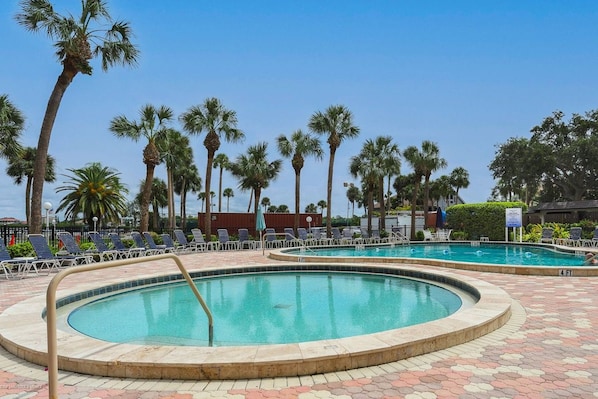 A pool area with two circular pools, lounge chairs, palm trees, and a clear sky.