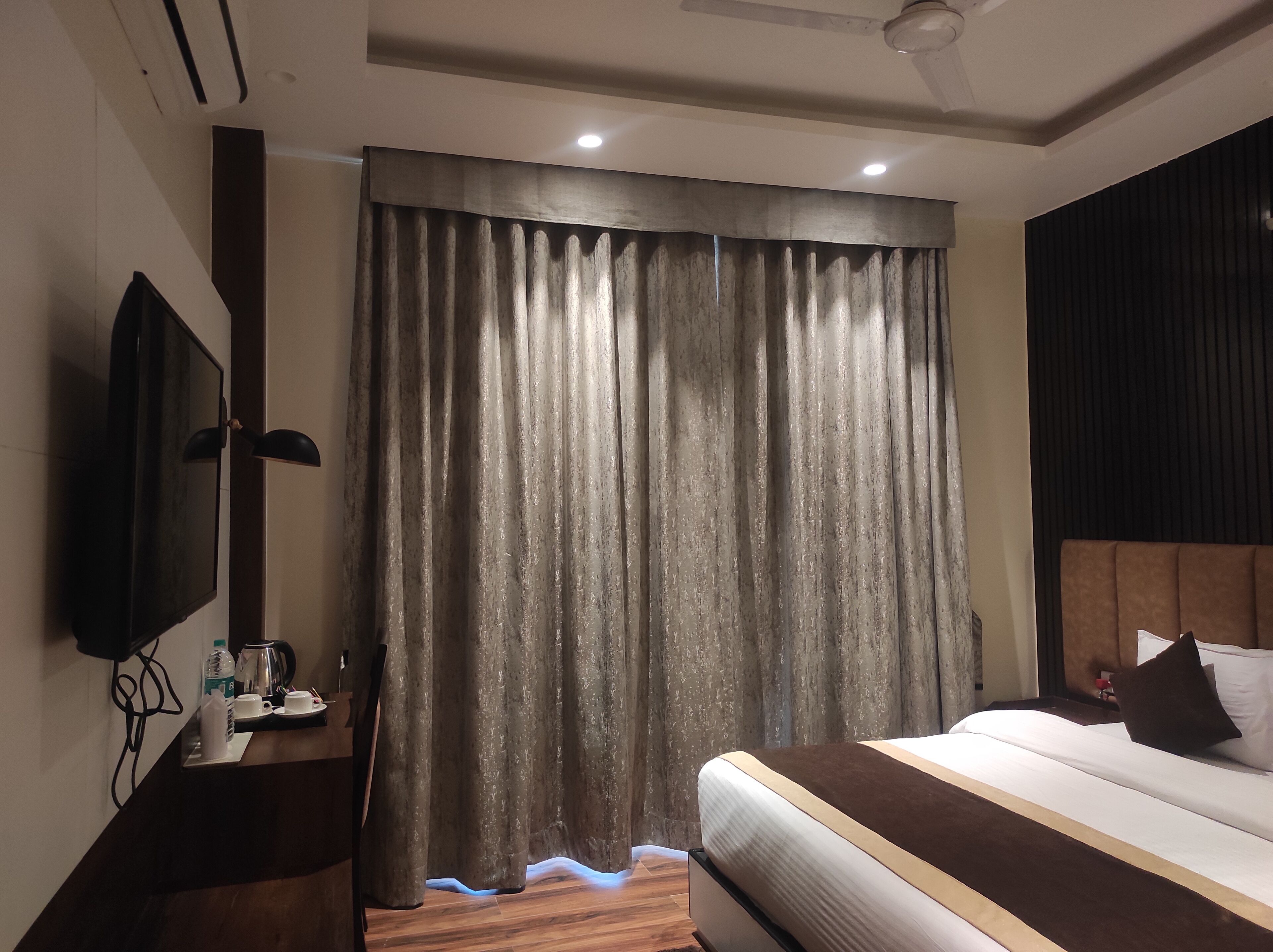 Habitación de hotel con cama, televisor de pantalla plana, escritorio con cafetera y ventana grande con cortinas.