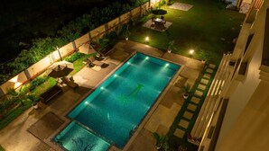 Piscina rectangular con una fuente de agua azul cristalina, rodeada por una zona de patio con mobiliario exterior y un jardín bien iluminado.