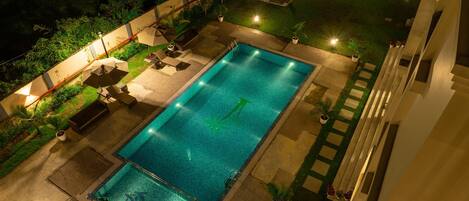 Piscina rectangular con una fuente de agua azul cristalina, rodeada por una zona de patio con mobiliario exterior y un jardín bien iluminado.