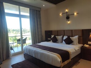 A hotel room with a large bed, a nightstand with a phone, a wall-mounted light fixture, and a view of the outdoors through a large window.