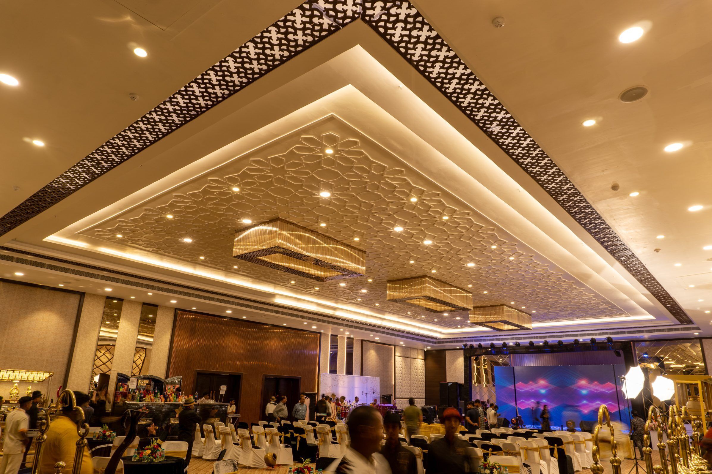 Un salón de banquetes con techo decorado, mesas dispuestas para un evento formal y un escenario con una pantalla grande.