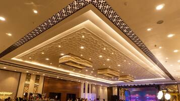 Un salón de banquetes con techo decorado, mesas dispuestas para un evento formal y un escenario con una pantalla grande.