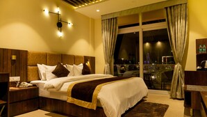 Una habitación de hotel moderna con una cama grande, un cabecero de madera, una mesita de noche con un teléfono y vistas a la ciudad a través de un ventanal con cortinas.