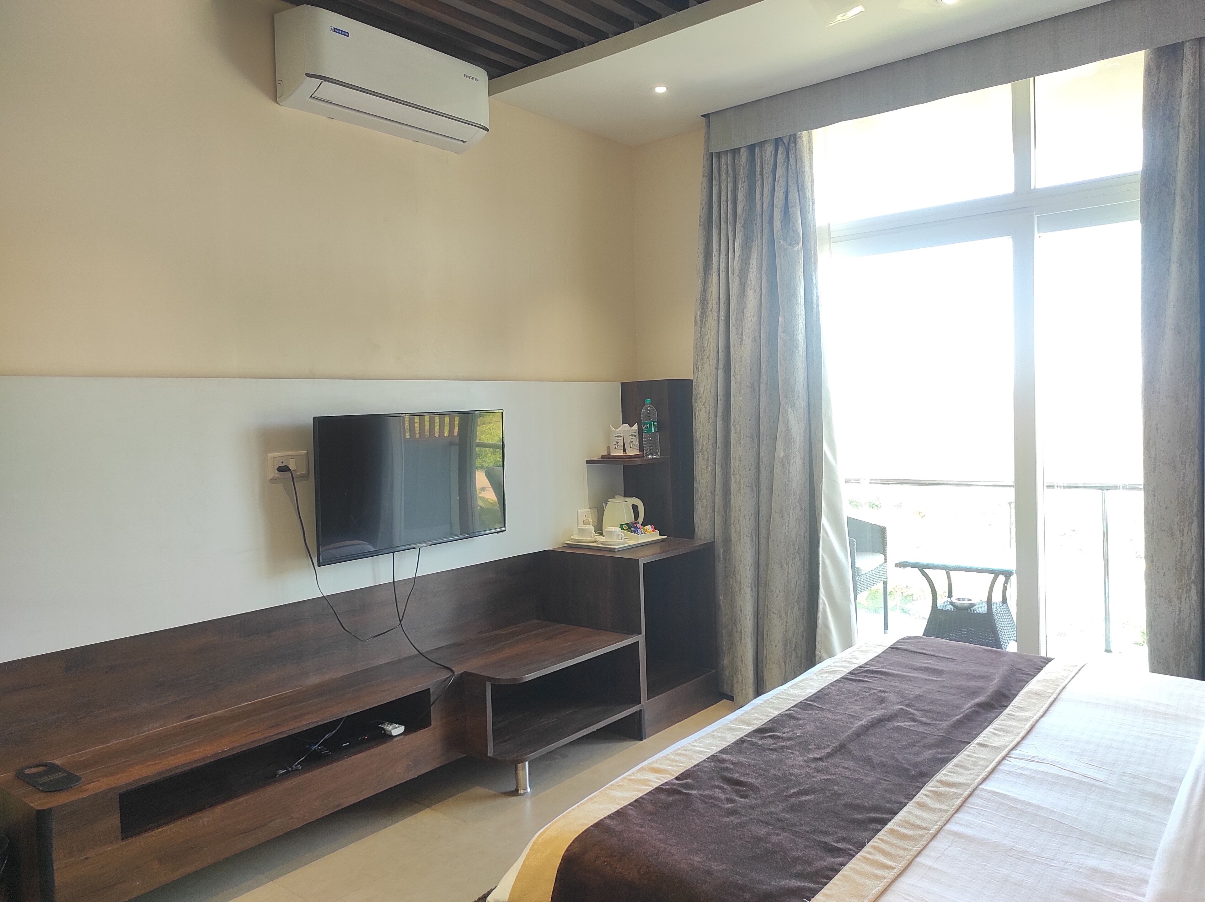 A hotel room with a bed, a TV mounted on the wall, a small table with a kettle and cups, a shelf with toiletries, and a window with curtains.