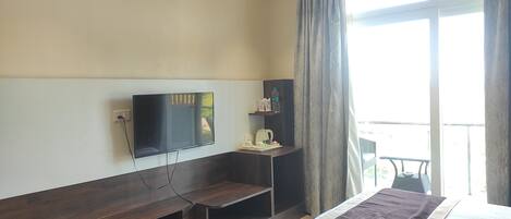 A hotel room with a bed, a TV mounted on the wall, a small table with a kettle and cups, a shelf with toiletries, and a window with curtains.