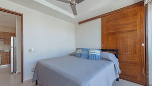 A bedroom with a bed, wooden wardrobe, ceiling fan, and a view into the kitchen.