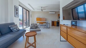 A modern living room with a sofa, a small wooden table, a kitchen area with a dining set, and a large TV.