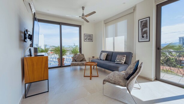 A modern living room with a sofa, two armchairs, a coffee table, and a TV stand. There is a large window with a view of buildings and palm trees.