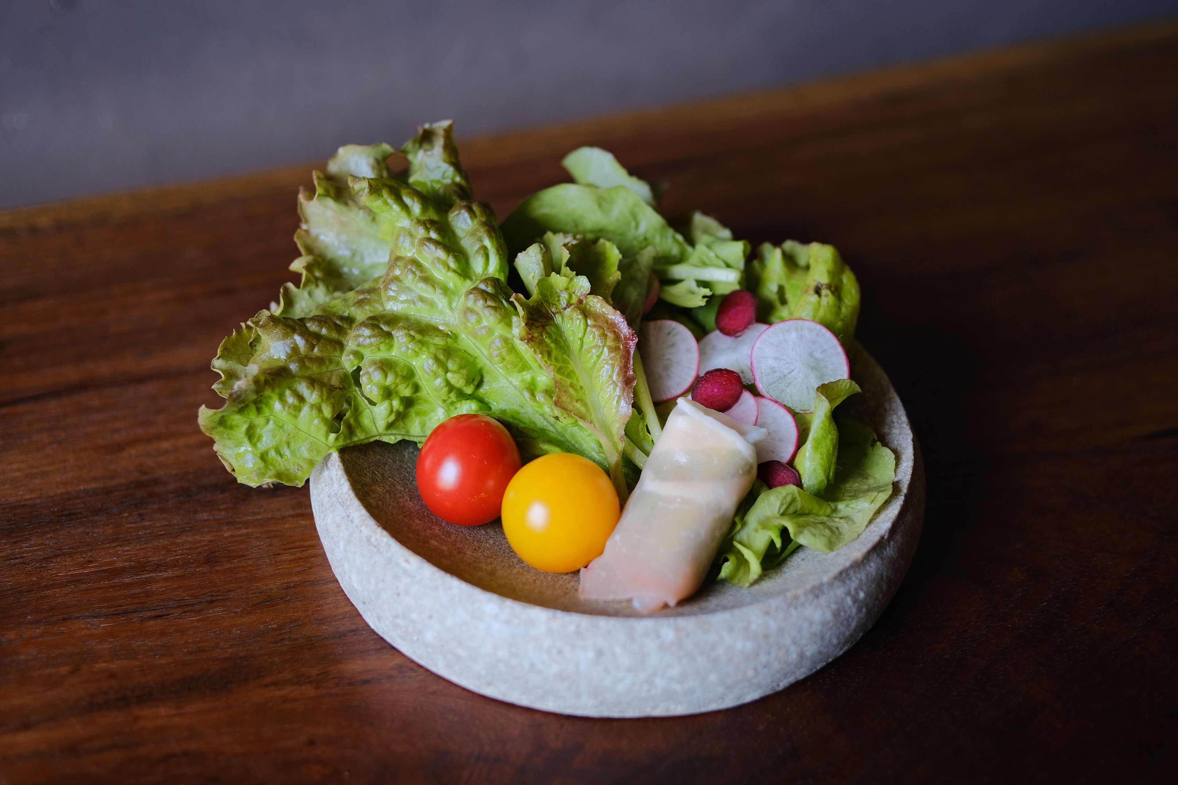 A salad with lettuce, radishes, a tomato, and a cheese slice on a round stone plate.