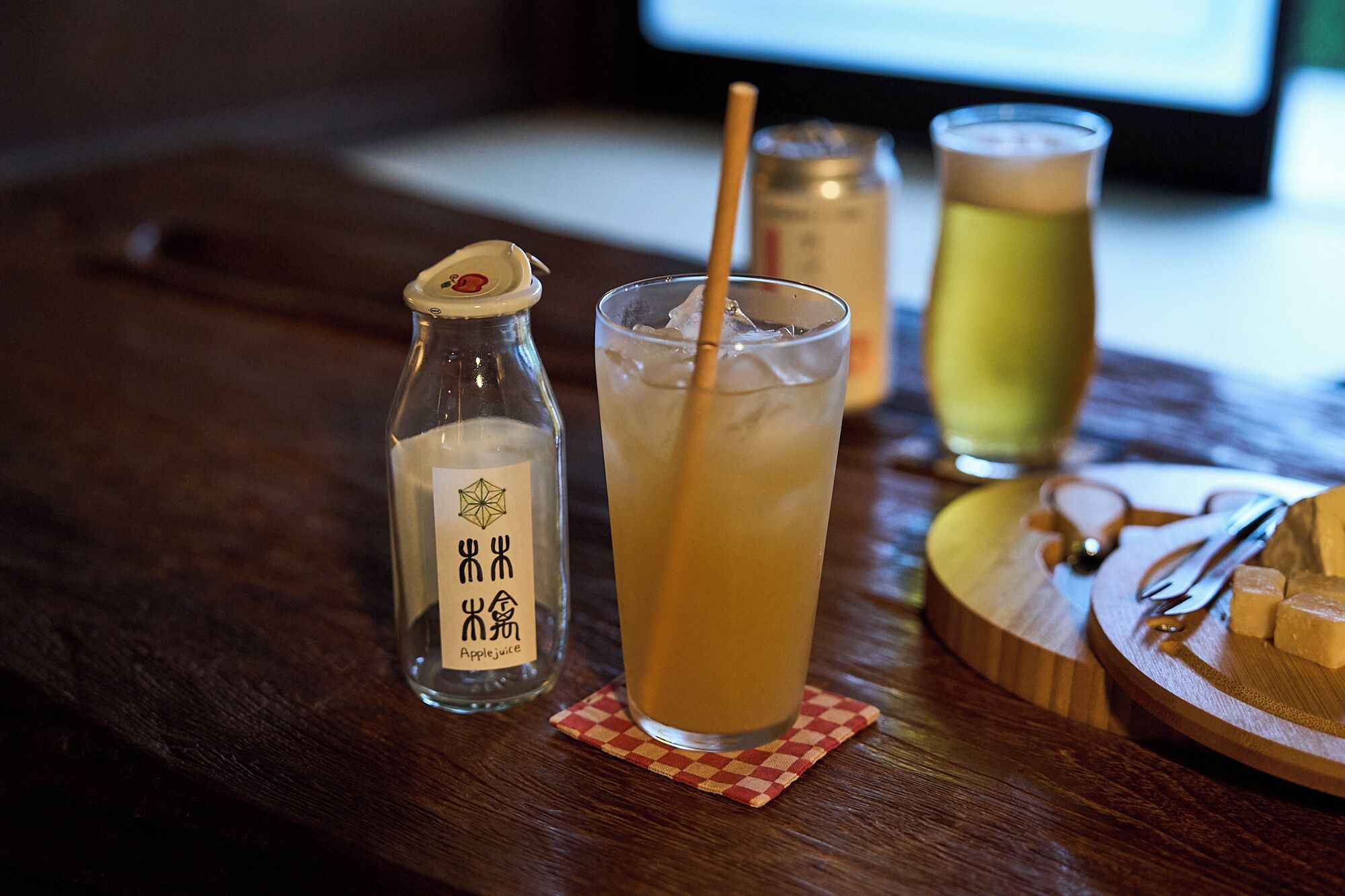 Un verre de jus de pomme avec une paille, une bouteille de jus de pomme et un verre contenant une autre boisson, le tout posé sur une table en bois.