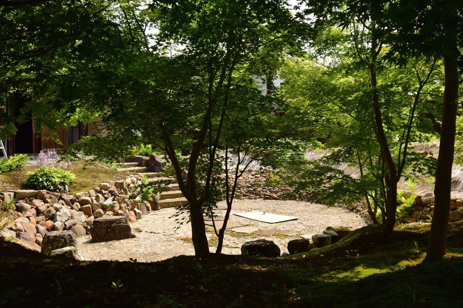 A traditional Japanese garden with stone pathways, a central stone platform, and surrounding trees.