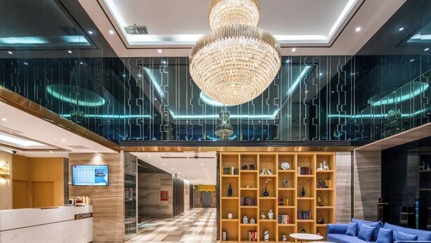 A modern lobby with a reception desk, a large chandelier, a blue sofa, and a bookshelf filled with books and decorative items. - YISHANG HOTEL (Guangzhou)