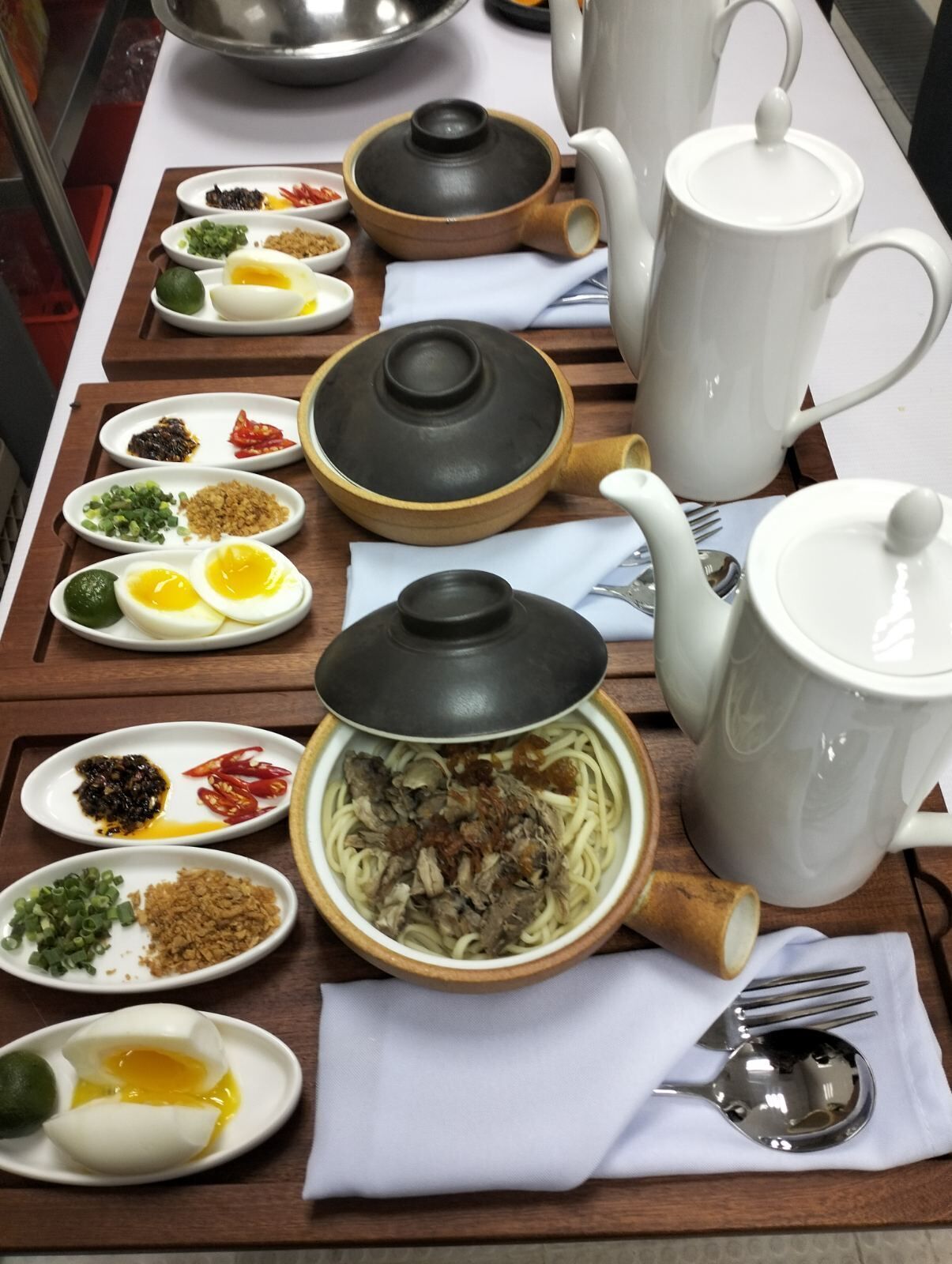 A dining table with various dishes, including noodles, eggs, and condiments. There are also teapots and a spoon on the table.