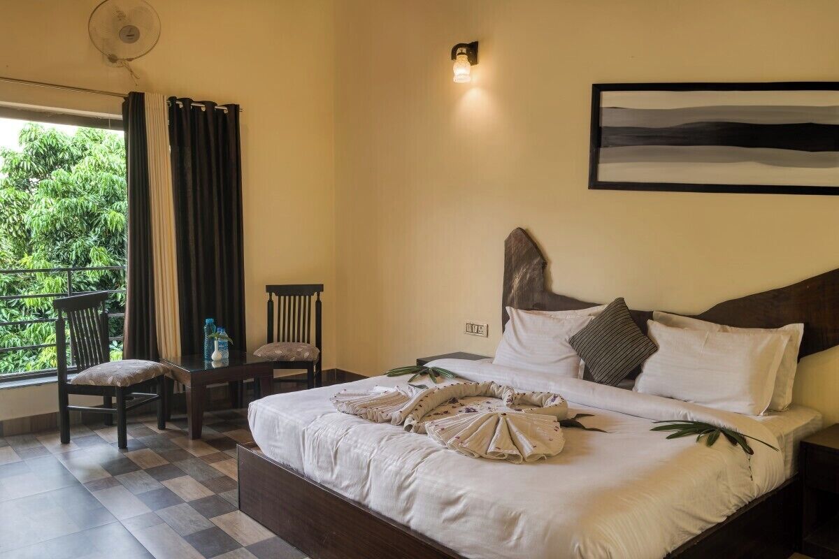 A hotel room with a large bed, a bedside table, a chair, a balcony with curtains, and a wall-mounted light fixture.