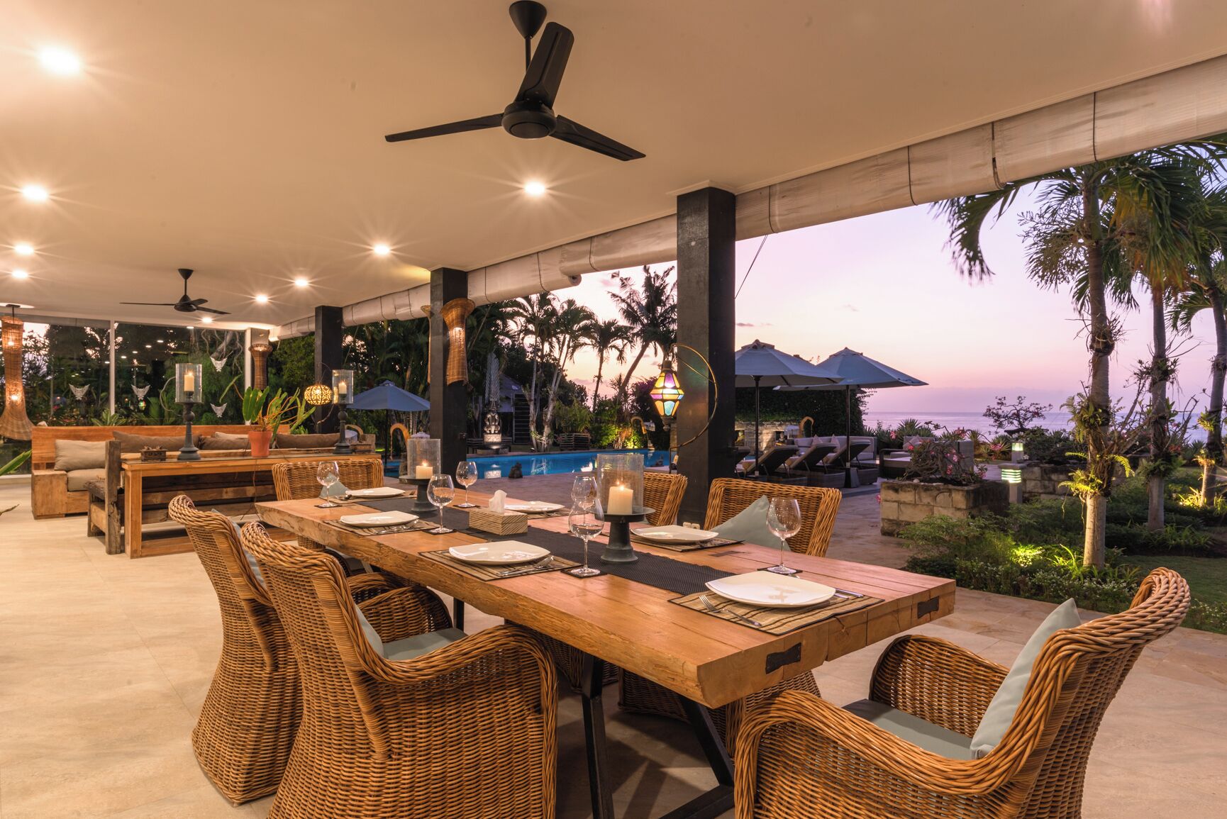 An outdoor dining area with wicker furniture, a pool, and a view of the ocean at sunset.