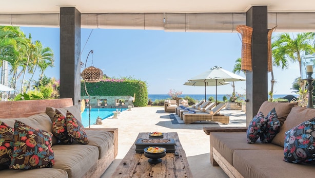 Zona de descanso junto a la piscina con mesa de madera, asientos acolchados y vista al océano.