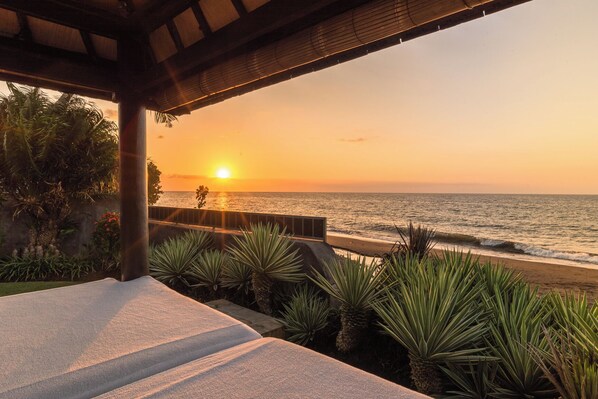 A beach view from a covered area with a railing, overlooking the ocean at sunset.
