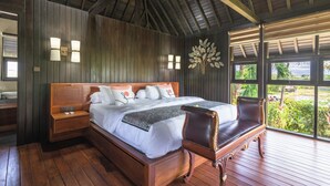 Un dormitorio amplio con una cama grande, cabecero de madera, mesitas de noche y vistas a abundante vegetación a través de amplias ventanas.