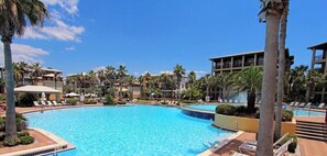 Ein großer Swimmingpool, umgeben von Palmen und Liegestühlen, mit blauem Himmel.