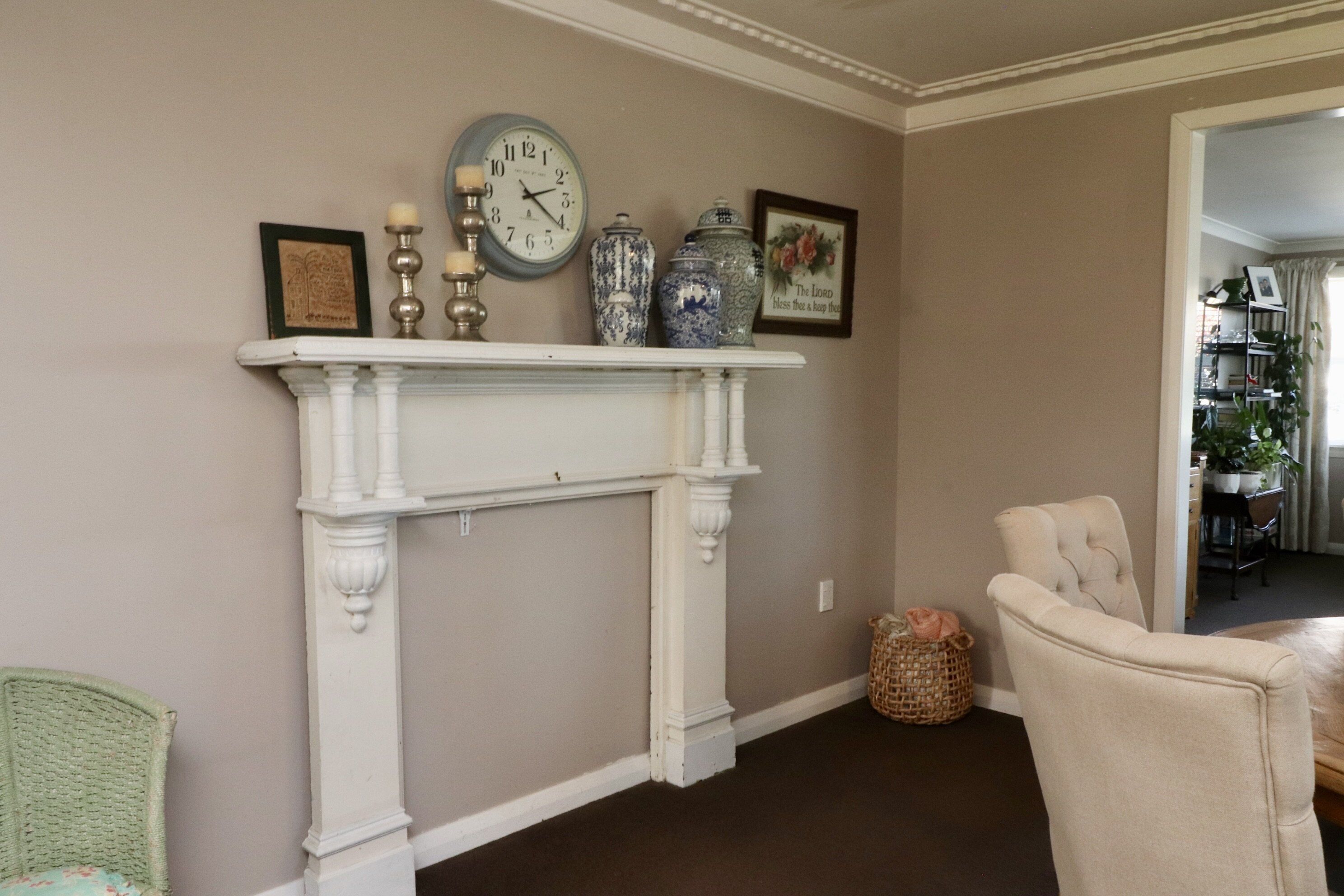 A living room with a white fireplace, a clock, and decorative vases. There is a wicker chair and a cushioned armchair.