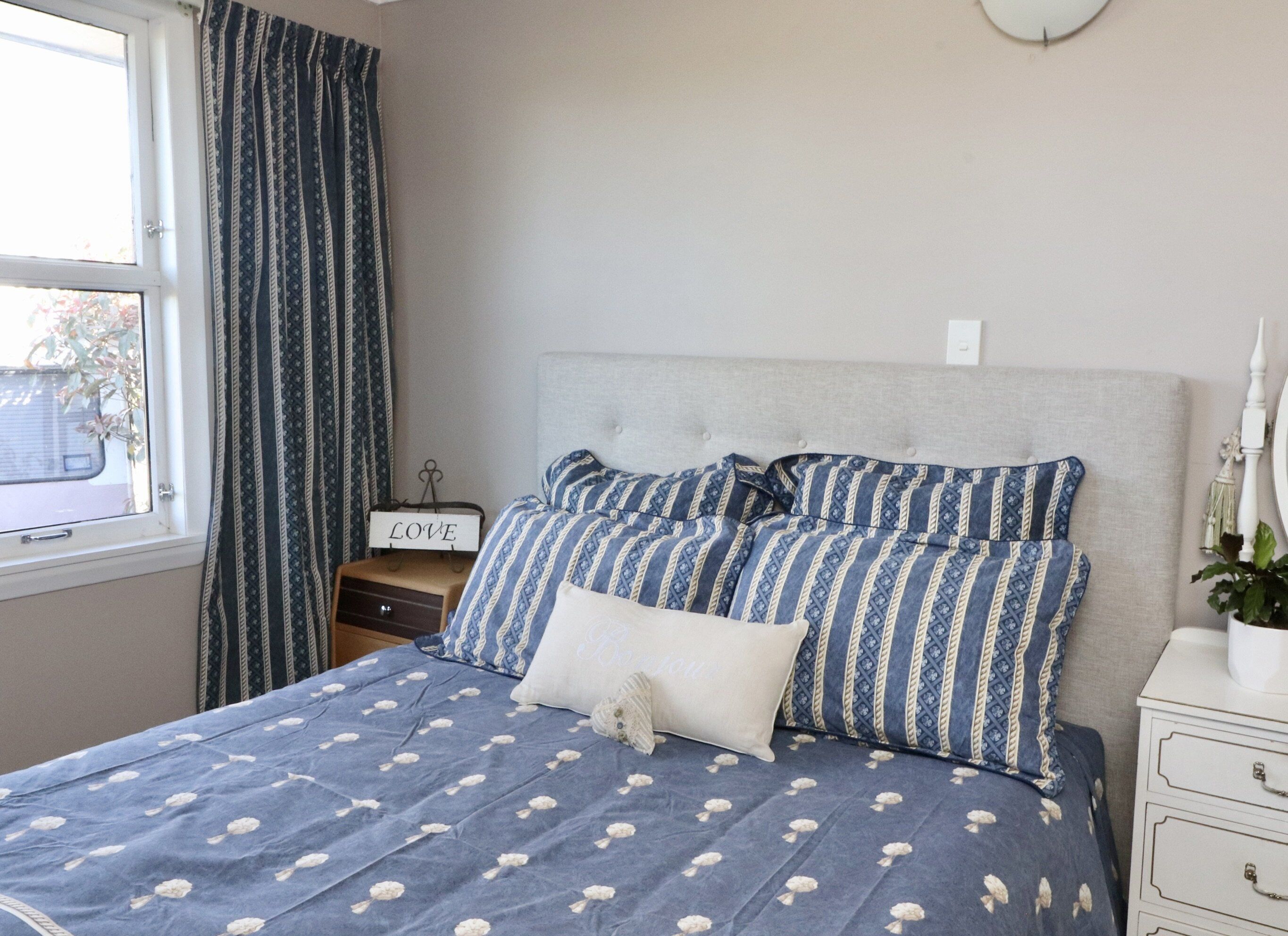 A bedroom with a bed, a bedside table, a nightstand, a window with curtains, and a wall clock.