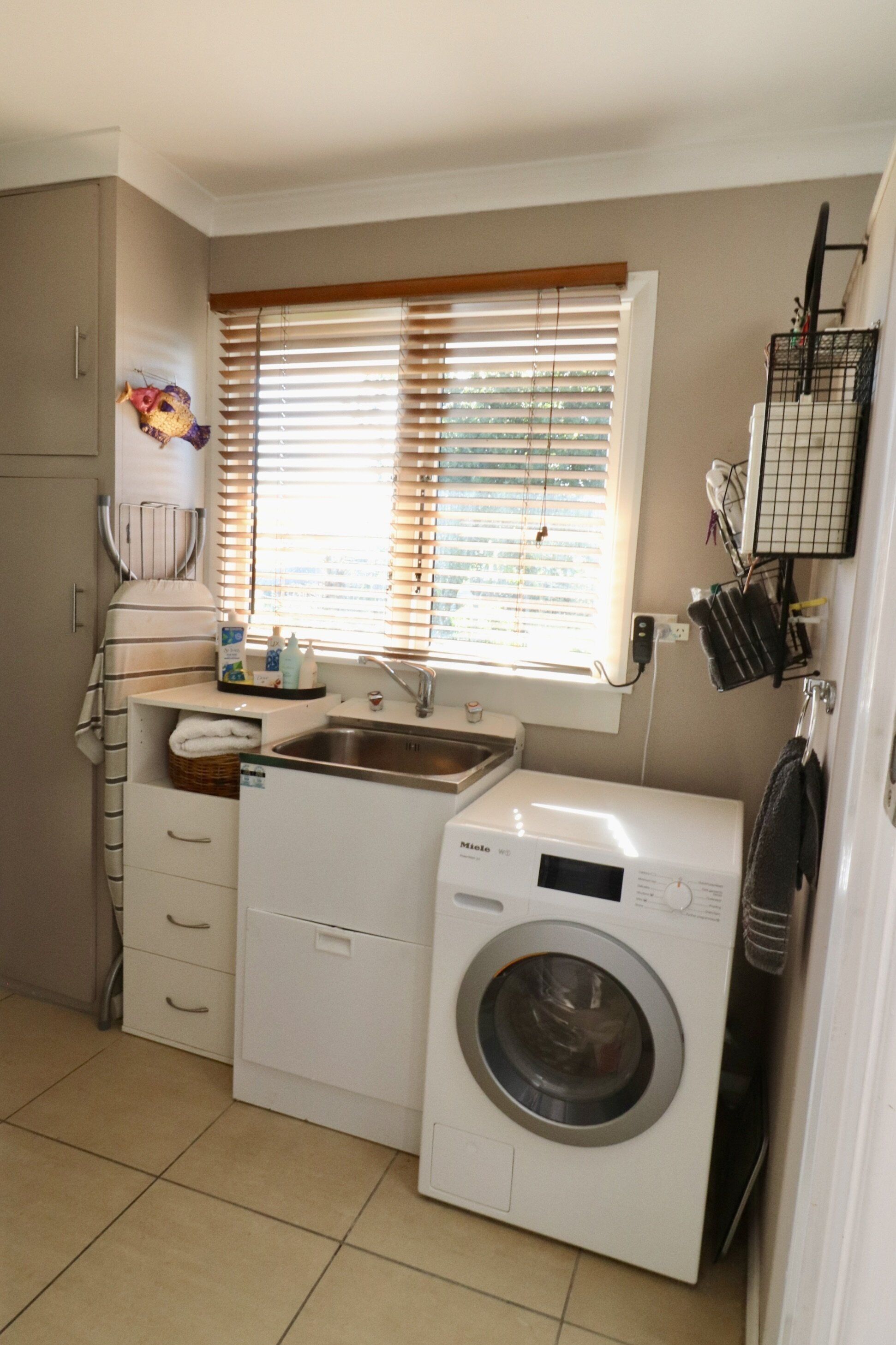 Un espace buanderie compact comprenant un lave-linge, un évier et une fenêtre avec des stores.