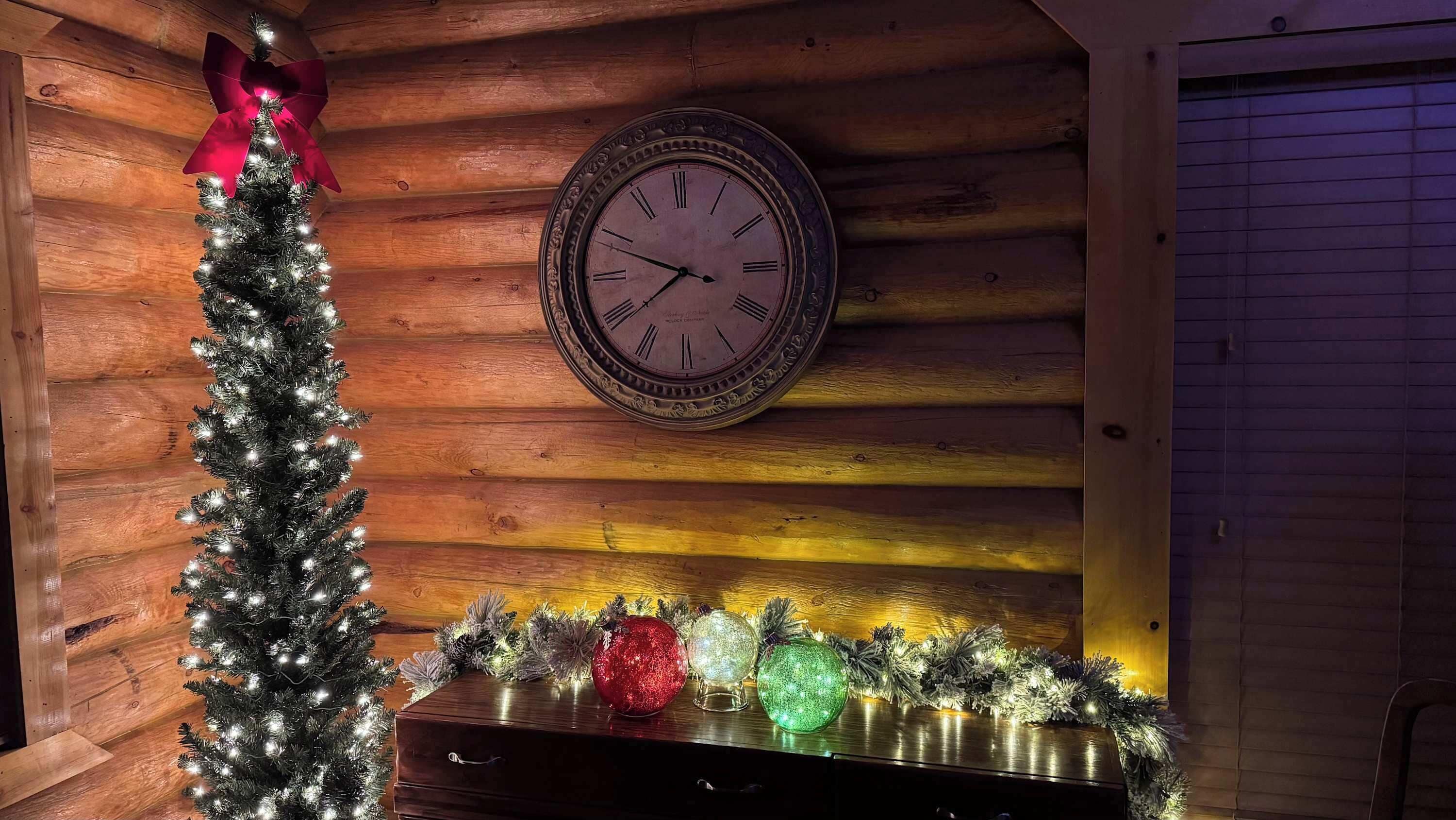 Interior de madeira com um relógio na parede, árvore de Natal decorada e exposição de enfeites coloridos.