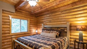 Une chambre située dans un chalet en rondins, avec un lit en bois, une table de chevet, une lampe et une fenêtre avec des stores.