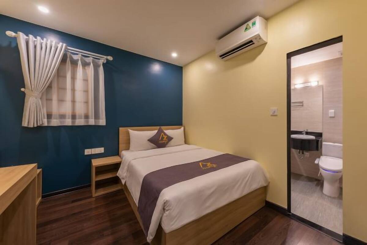 A hotel room with a bed, a small bedside table, a window with curtains, an air conditioning unit, and a bathroom visible through an open door.