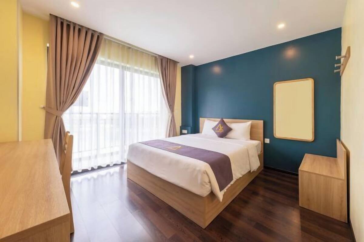 A hotel room with a large bed, a wooden desk, a nightstand, and a window with curtains.