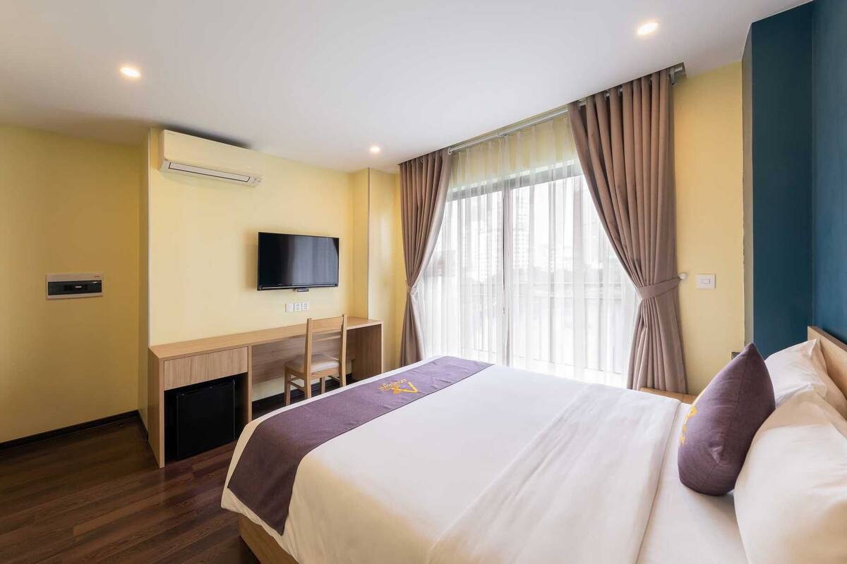 A hotel room with a bed, a desk with a chair, a television, and a window with curtains.
