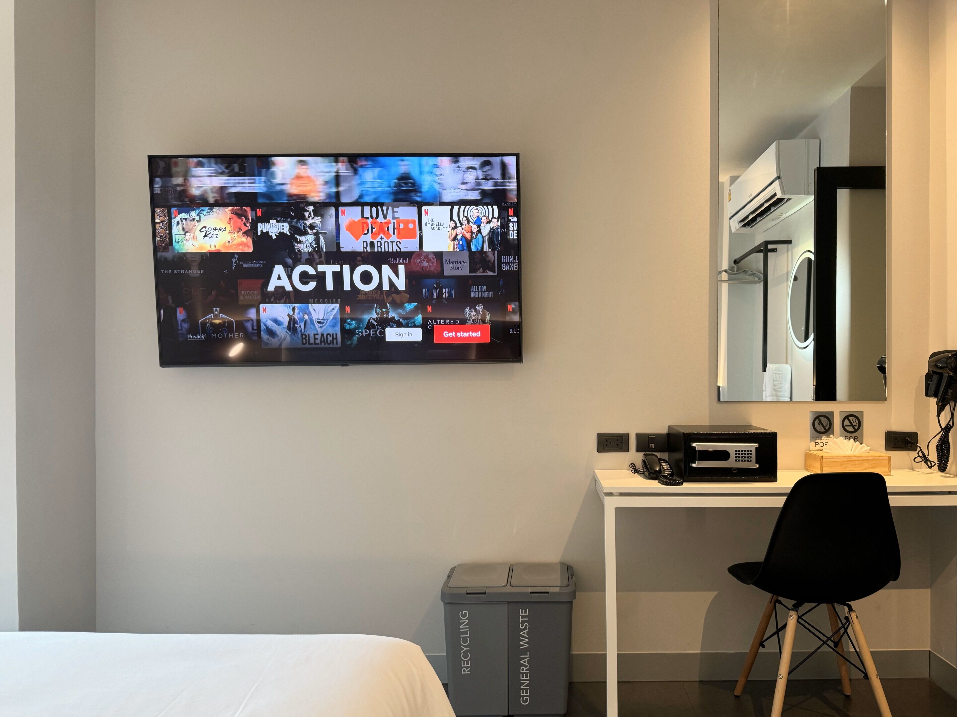 Hotellihuone, jossa on suuri televisio, jolta voi katsoa useita suoratoistopalveluita, valkoinen työpöytä, jossa on tietokone ja puhelin, musta tuoli ja kierrätysastia, jossa on merkintä ”YLEINEN JÄTE”.
