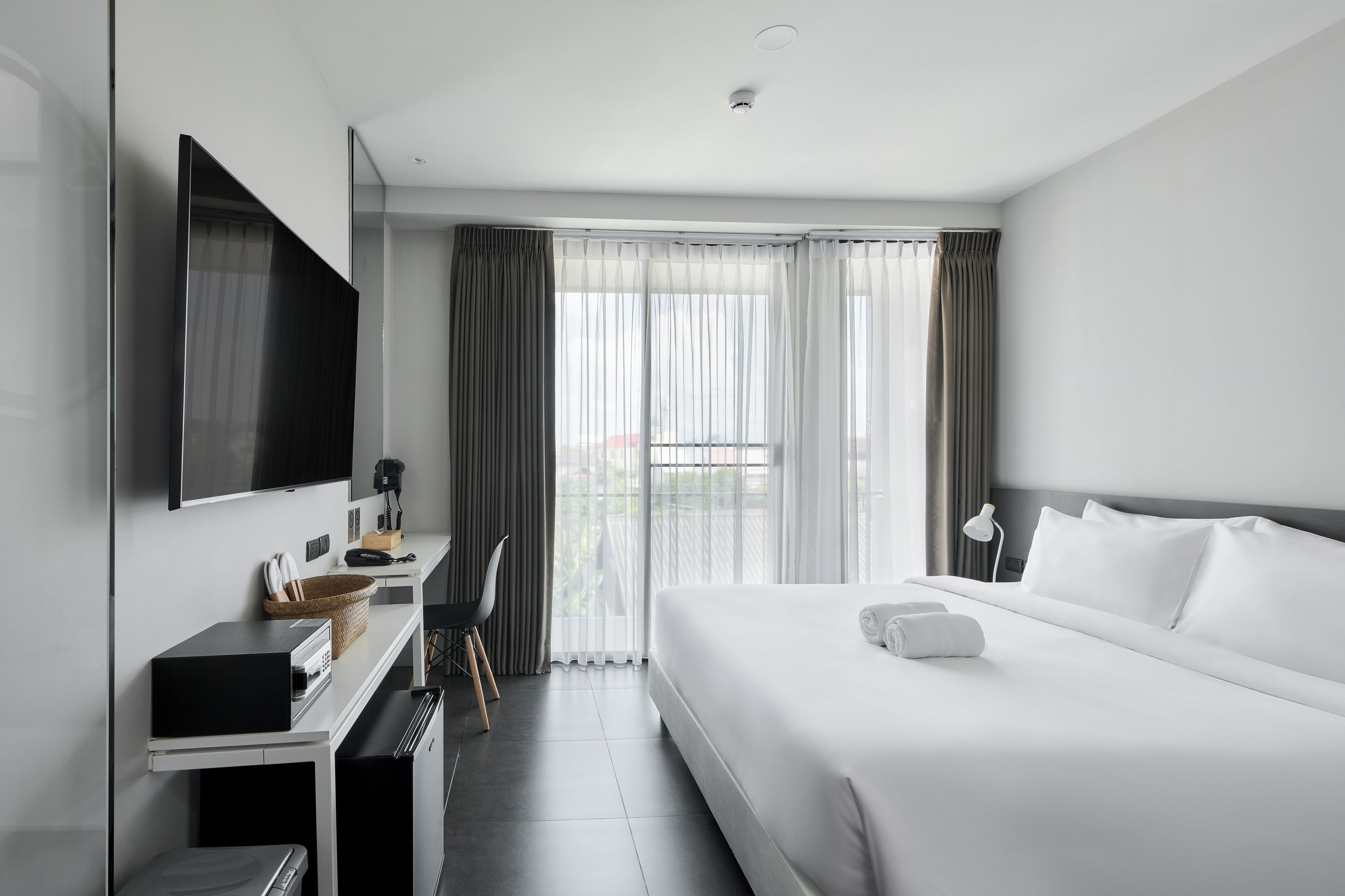 A modern hotel room with a large bed, a flat-screen TV, a desk with a computer, and a view of the outdoors through sheer curtains.