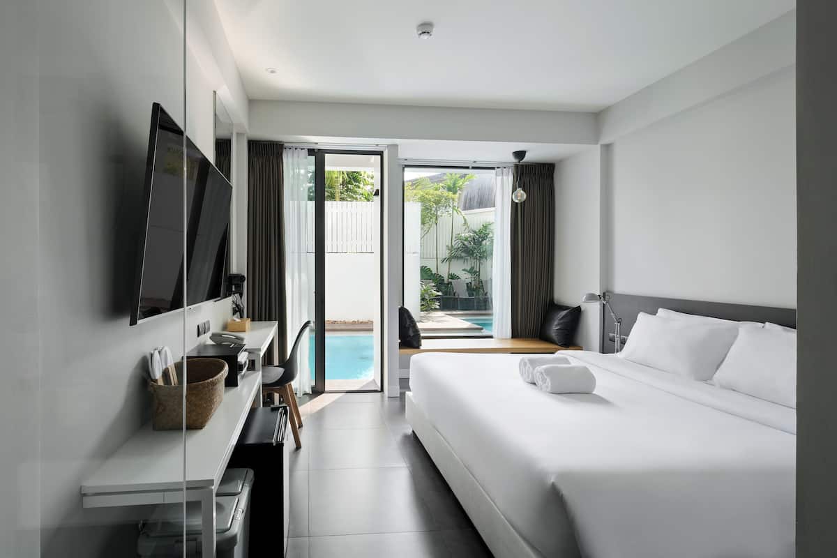 A modern hotel room with a large bed, a desk with a computer, and a view of a swimming pool and garden.