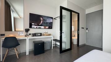 Habitación de hotel moderna con cama, escritorio con computadora, televisor y baño visible a través de un vidrio.