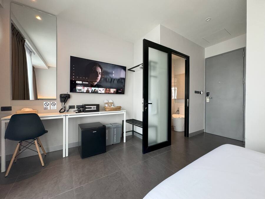 Habitación de hotel moderna con cama, escritorio con computadora, televisor y baño visible a través de un vidrio.