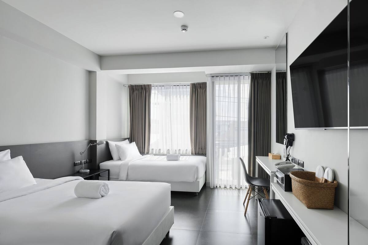 A modern hotel room with two beds, a desk, a TV, and a large window with curtains.