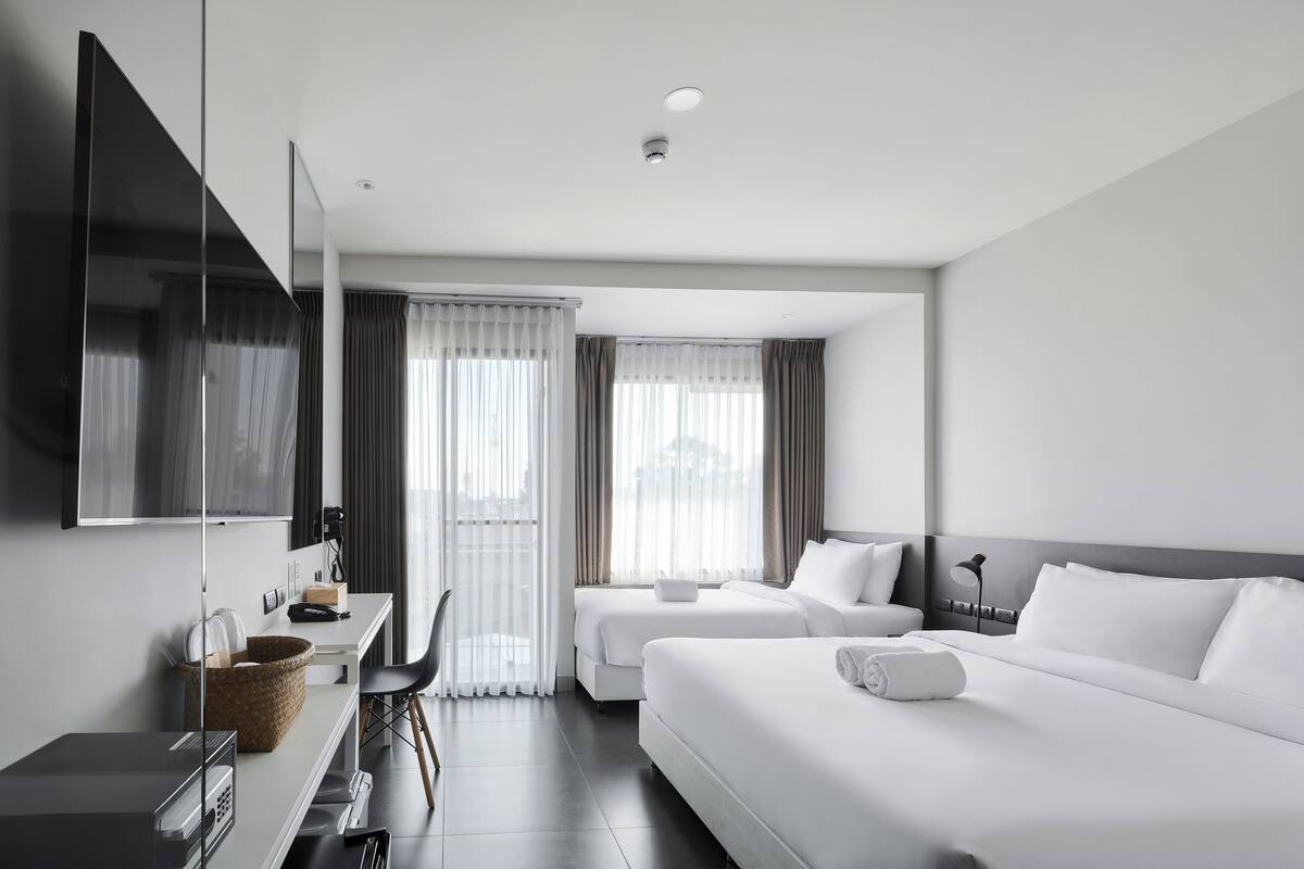 A modern hotel room with a flat-screen TV, a desk with a computer, two beds, and a large window with curtains.