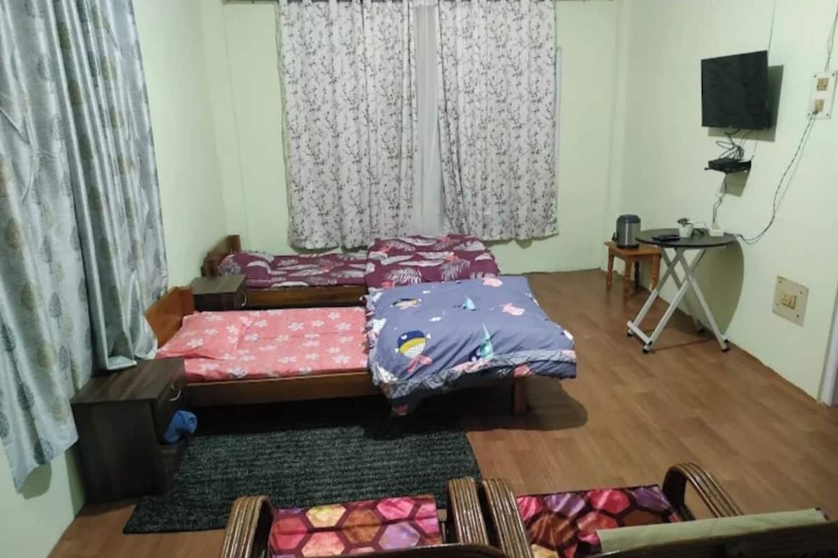 A bedroom with two single beds, a small table with a chair, a TV mounted on the wall, and a window with curtains.