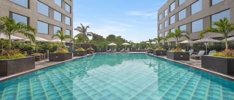 A swimming pool with a grid pattern, surrounded by lounge chairs and umbrellas, with a modern building in the background.
