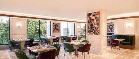 A modern dining area with multiple tables and chairs, large windows offering a view of the outdoors, and a central column with artwork.