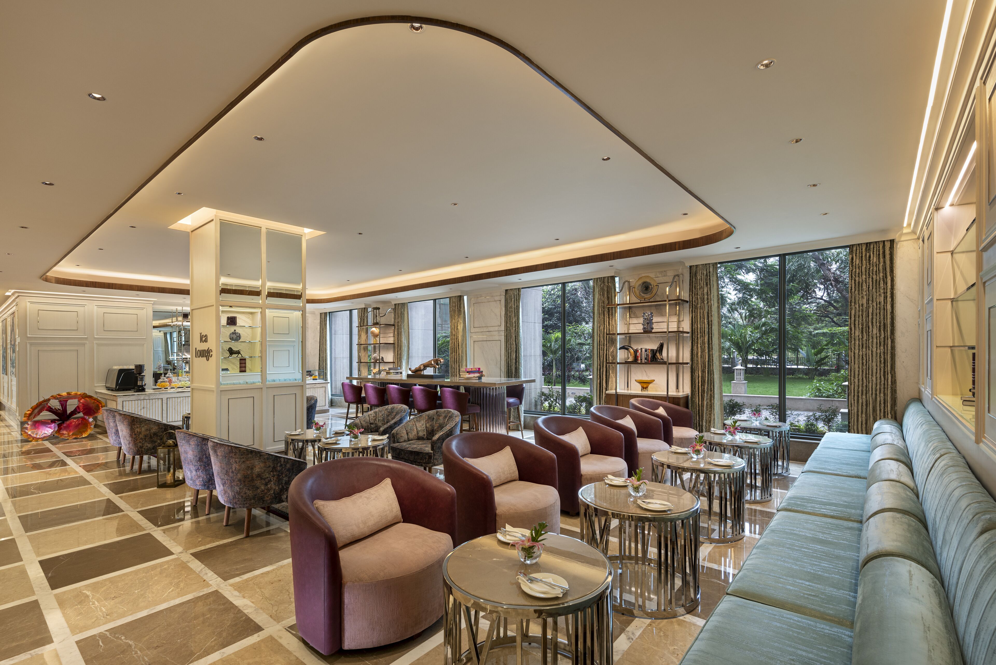 A spacious lounge area with multiple seating options including sofas and armchairs, a bar area with a counter, and a view of a garden through large windows.