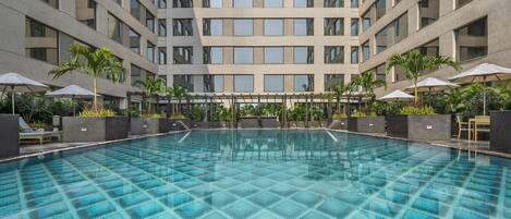 A large indoor swimming pool with a tiled floor, surrounded by lounge chairs and umbrellas, and a high-rise building in the background.